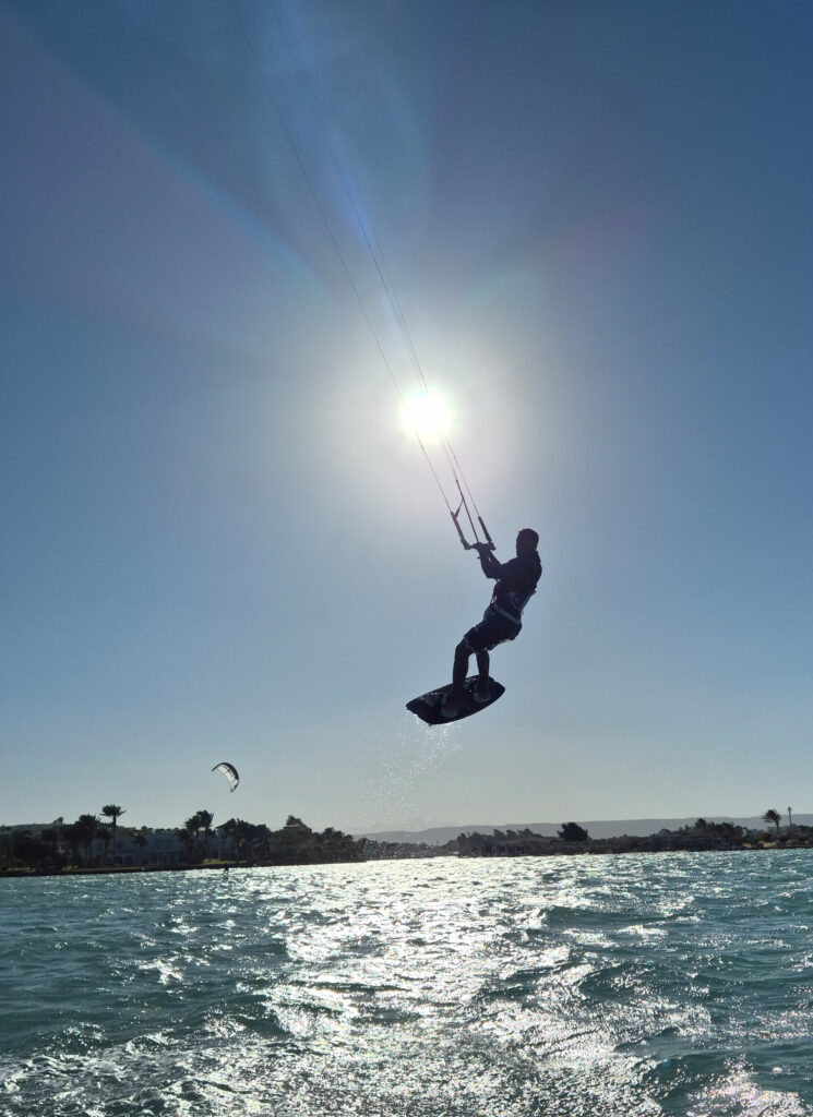 Kitesurfer jumping above the water, suspended by the kite, with the sun in the background and the sea below. Kitesurfer springt über dem Wasser, gehalten vom Kite, mit Sonne im Hintergrund und Meer darunter.