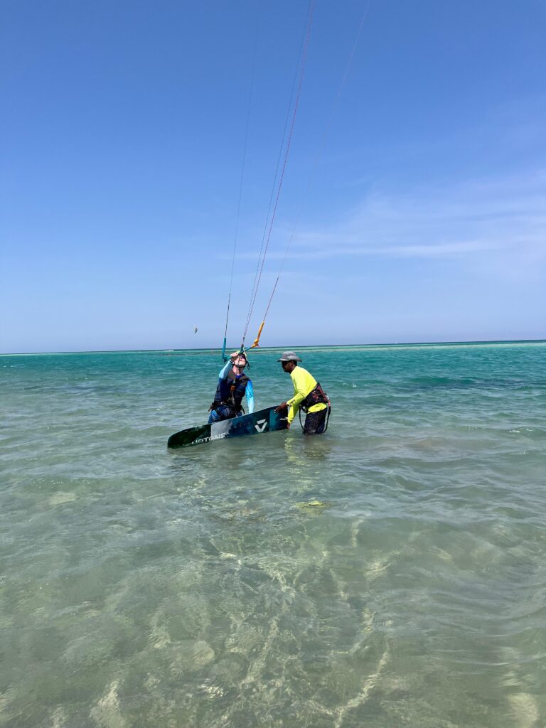 Two people standing knee-deep in shallow sea water, one holding a kitesurf board while both prepare the kite. Zwei Personen stehen knietief im flachen Meerwasser, eine Person hält ein Kitesurfboard, während beide den Kite vorbereiten.