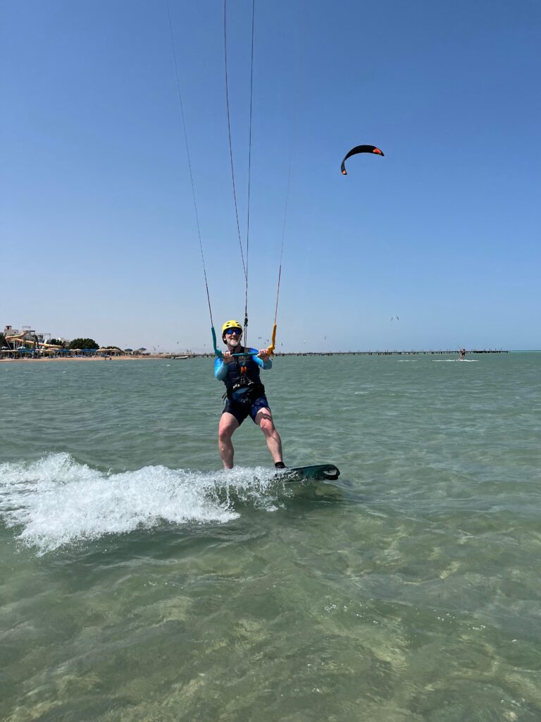 Kitesurfer riding on a board in shallow sea water while holding the kite control bar, with the beach and additional kites in the background. Kitesurfer fährt im flachen Meerwasser auf einem Board und hält die Kite-Controlbar, im Hintergrund Strand und weitere Kites.