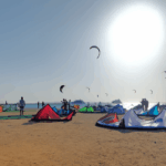 Several kites lying on a sandy beach, with people standing nearby and kites flying over the sea in the background. Mehrere Kites liegen am Sandstrand, im Hintergrund stehen Personen und fliegen Kites über dem Meer.