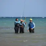 Three people standing knee-deep in the sea, jointly holding a kite control bar while preparing the kite. Drei Personen stehen knietief im Meerwasser und halten gemeinsam eine Kite-Controlbar, während sie den Kite vorbereiten.