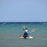 Three people standing knee-deep in the sea, jointly holding a kite control bar while preparing the kite. Drei Personen stehen knietief im Meerwasser und halten gemeinsam eine Kite-Controlbar, während sie den Kite vorbereiten.