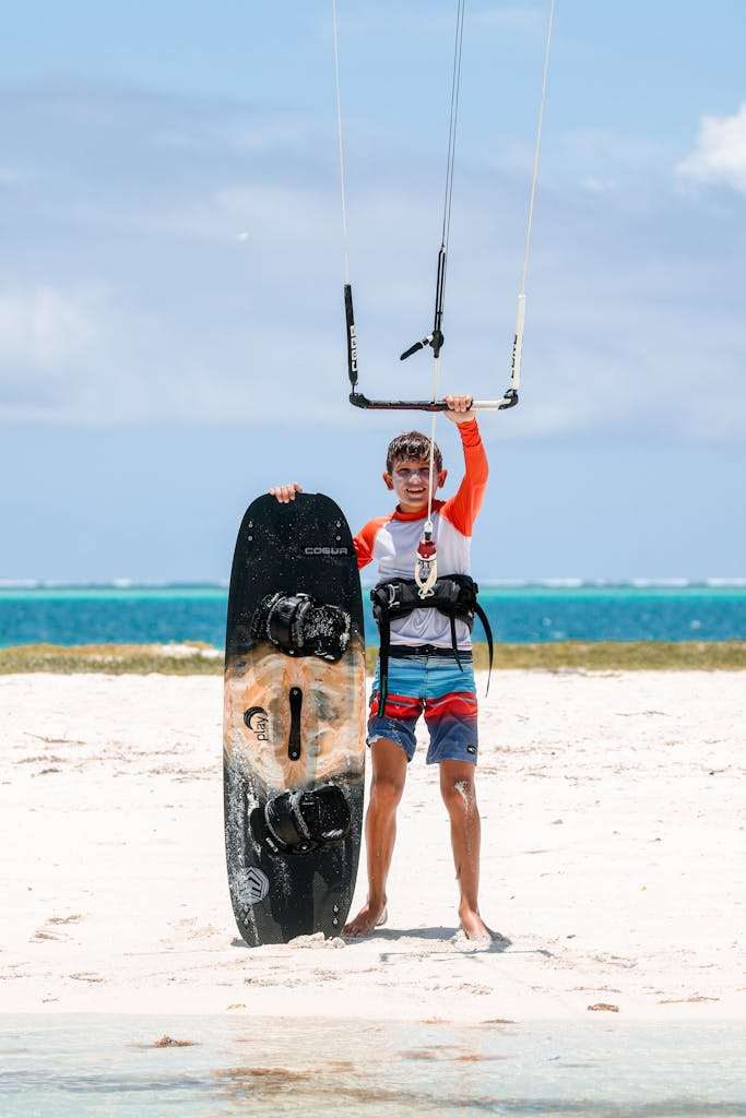 A boy on the beach holding a kite and wakeboard, ready for kitesurfing under a clear blue sky.